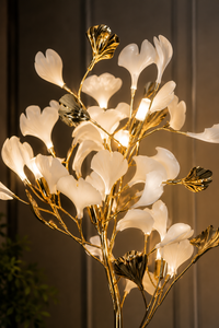 A modern tree-style floor lamp with illuminated white magnolia-like flower petals and metallic gold branches, shown against a dark paneled background.