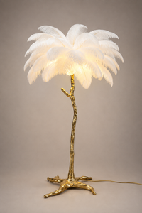 A palm tree table lamp with a glowing white feathered canopy and a textured metallic gold trunk and base, photographed against a neutral warm gray studio background.