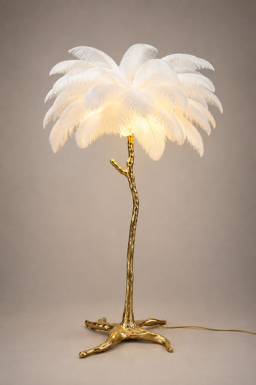 A palm tree table lamp with a glowing white feathered canopy and a textured metallic gold trunk and base, photographed against a neutral warm gray studio background.