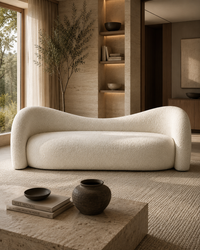 A modern curved lounge sofa upholstered in textured cream bouclé fabric, set in a warm, minimalist interior with a stone coffee table, woven rug, and built-in shelving.
