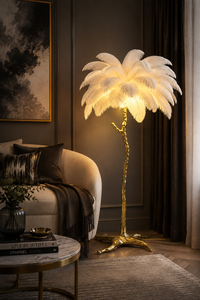 A luxury floor lamp styled in a modern living room. The lamp features a glowing white feathered palm-style shade on a twisted gold metal trunk and base, set next to a beige armchair and a dark curtain.