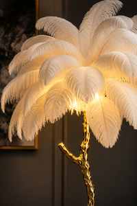A decorative floor lamp with a large cluster of white ostrich feathers surrounding a glowing warm light, mounted on a textured metallic gold branch-shaped stand.