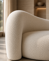 Close-up of a modern curved armchair upholstered in textured cream bouclé fabric, set in a softly lit interior with warm neutral walls and a window in the background.