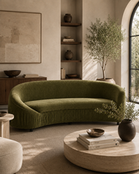 A curved olive green velvet sofa with fringe trim in a warm, modern living room setting with a round wooden coffee table and neutral decor.