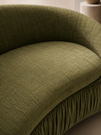 Close-up of an olive green upholstered armchair with a curved back, textured woven fabric, and a pleated skirt base.