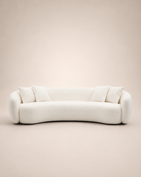 A modern curved sofa upholstered in a soft off-white bouclé fabric, featuring four matching throw pillows, set against a solid warm beige background.