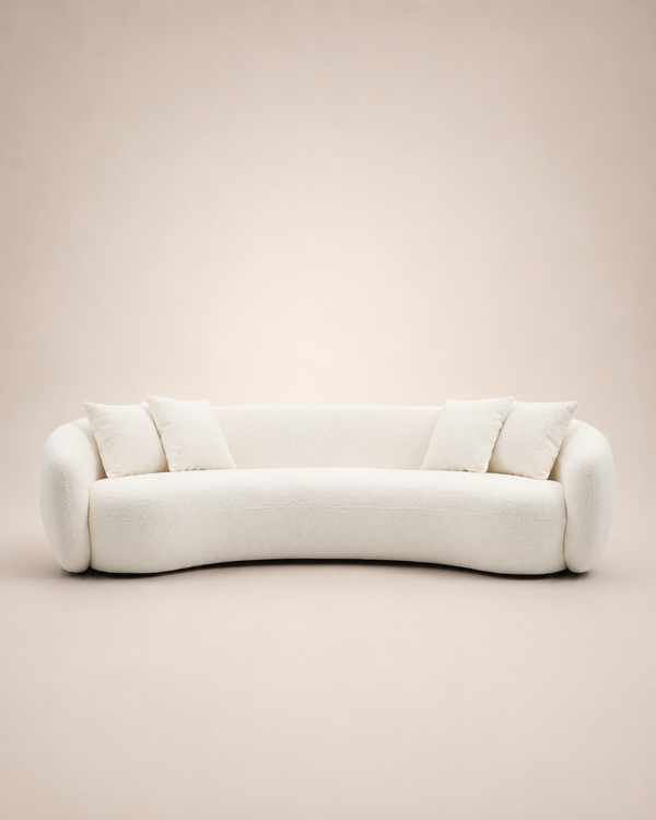 A modern curved sofa upholstered in a soft off-white bouclé fabric, featuring four matching throw pillows, set against a solid warm beige background.
