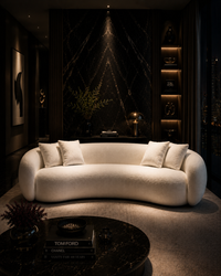 A modern curved white boucle sofa with four matching throw pillows, set in a luxurious, dimly lit contemporary living room with a dark marble accent wall and built-in shelves.
