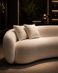 A modern curved sofa upholstered in a textured cream bouclé fabric, styled with two matching throw pillows in a dark, elegantly lit living room.