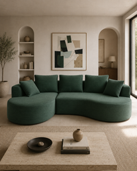 A curved, modular upholstered sofa in deep forest green sits in a modern, warm-toned living room with beige walls, built-in shelves, and a low stone coffee table.