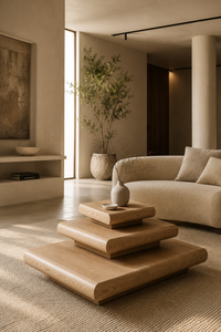 A minimalist three-tier wooden coffee table in a warm, modern living room setting with a beige sofa, textured rug, and a potted plant in the background.