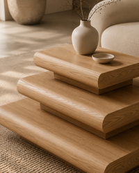 Three-tier wooden side table with rounded edges in a warm light oak finish, styled with a small ceramic vase and dish in a softly lit interior setting.