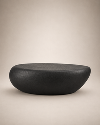 A modern, minimalist oval-shaped black stone or concrete stool with a textured matte finish, set against a warm beige gradient background.