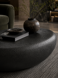 A dark textured oval coffee table in a modern living room setting, styled with a book, small bowl, and a rustic ceramic vase containing dried branches.