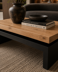 A modern low coffee table with a light oak wood top and black metal base, styled in a living room setting with a dark ceramic vase, books, and a bowl on top.