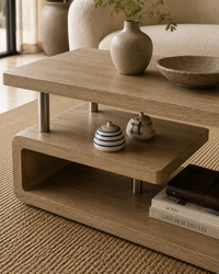 A modern two-tier wooden coffee table with a warm oak finish and slim metal legs, styled in a cozy living room setting with decorative ceramic vases, books, and a bowl.