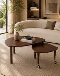 Two nested oval wooden coffee tables in a warm, modern living room. The larger table is dark walnut brown with tapered legs, and the smaller matching table has caster wheels.