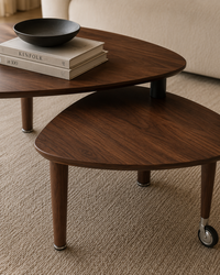 Two overlapping triangular wooden nesting tables with dark walnut-like grain, shown in a styled interior. The front table has small casters at the base and holds a black bowl and stacked books on top.