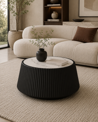 A modern round coffee table with a black pleated base and a white marble top, styled in a contemporary living room with a cream bouclé sofa and a brown accent pillow.