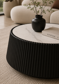 A round black coffee table with a fluted lower body and a white marble top, styled in a modern living room with a black vase and a book on top.