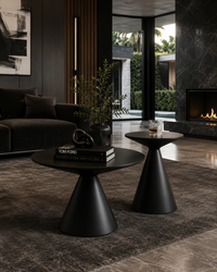 Two modern black tapered side tables in a stylish, dark-themed living room. One larger round table holds a book and a small plant, while a smaller matching table sits beside it with a glass of whiskey.