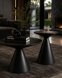 Two matte black modern side tables in a stylish, dark-themed living room. The larger round coffee table on the left holds a black vase with greenery, a decorative black ring, and a stack of books, while the smaller matching side table on the right holds a cut glass with an amber drink.