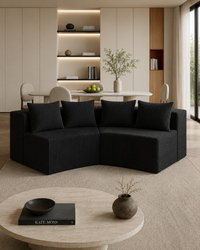 A modern curved black sectional sofa with ribbed upholstery in a minimalist beige living room, with a round coffee table, built-in shelving, and dining area in the background.