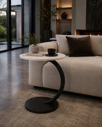 A modern round side table with a white marble top and a curved black metal base, styled in a contemporary living room next to a beige sofa.