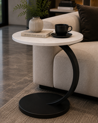 A modern round side table with a white marble top and a curved black metal base, styled next to a beige sofa with a black coffee cup and a small vase on top.