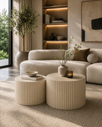 A modern living room scene featuring two ribbed cylindrical coffee tables in a warm cream color, styled with vases and books in front of a beige sectional sofa.