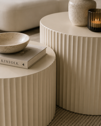 Two round ribbed side tables in a warm cream tone, styled with a stone bowl, a book, and a candle in a softly lit interior setting.