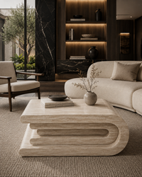 A modern curved coffee table in a light beige stone or wood finish, styled in a luxurious contemporary living room with a cream sofa, a mid-century armchair, and a dark wood shelving unit in the background.