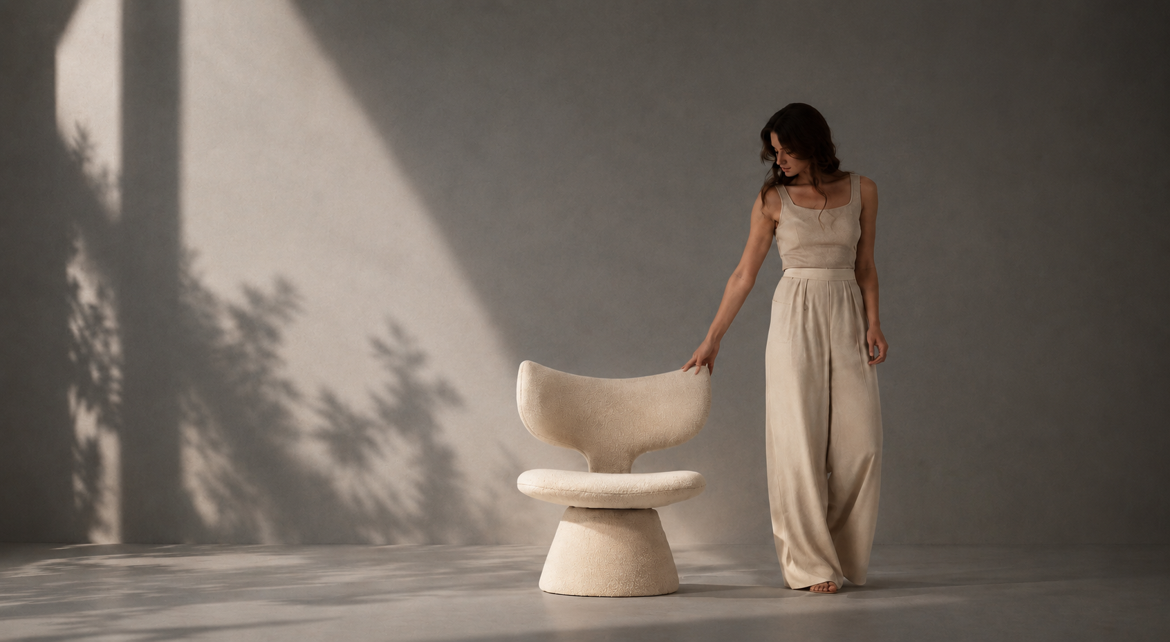 A woman in a beige sleeveless jumpsuit stands beside a sculptural molded chair in a minimalist studio with dramatic shadows cast across a gray wall and floor.